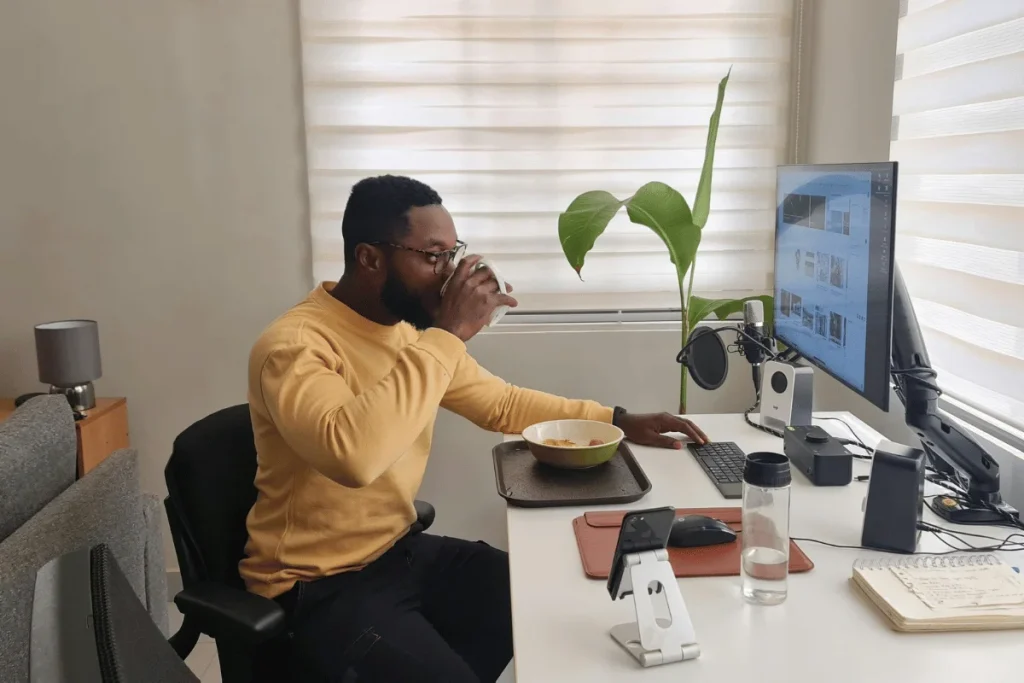 Man working at his home office desk, sipping coffee and browsing on a desktop monitor.