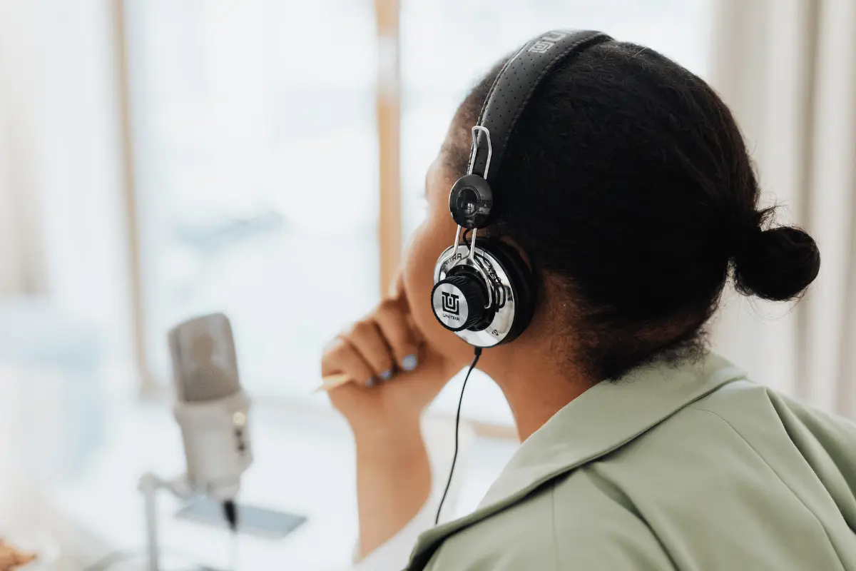 A woman wearing over-ear headphones sits beside a studio microphone, resting her chin on her hand as she looks toward a bright window, appearing thoughtful and focused.