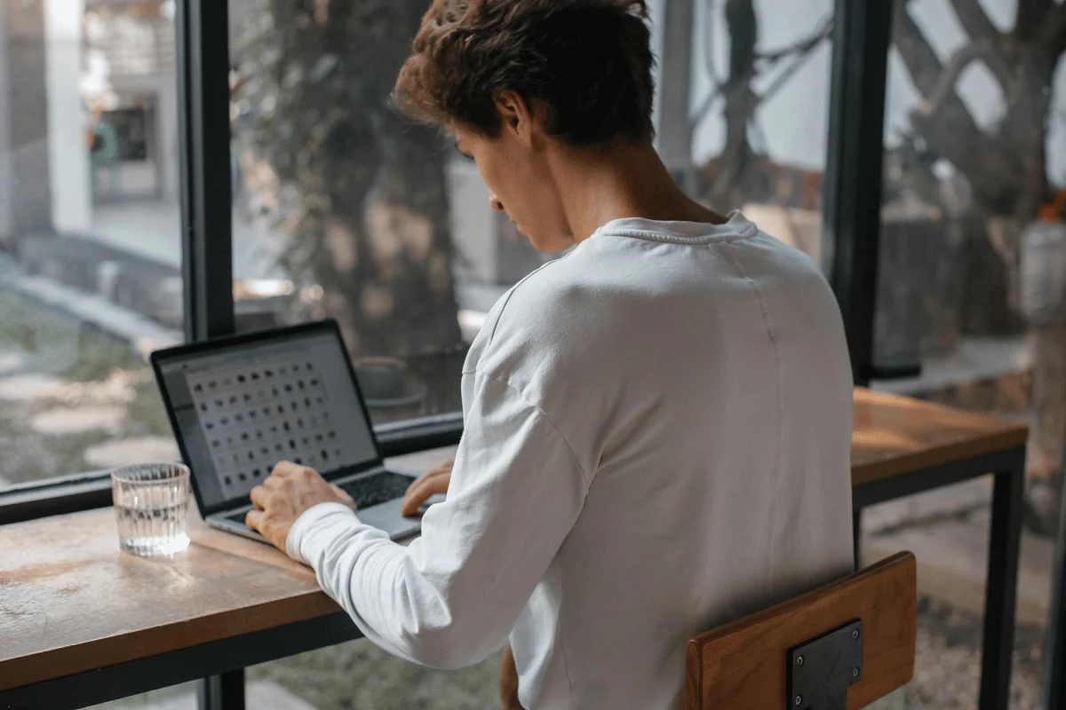 Man in a white shirt working on a laptop at a wooden desk near a window.