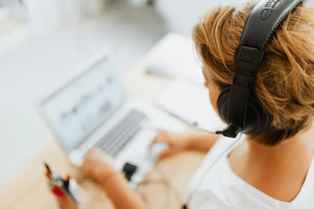 A woman with short hair wears headphones while typing on a laptop, surrounded by stationery on a light-colored desk.