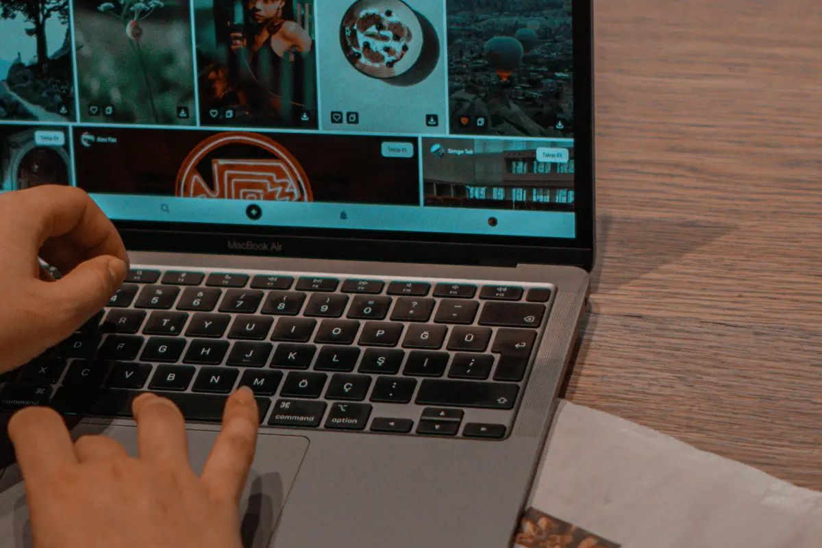 Hands type on a MacBook showing a dark, tiled photo gallery with food and travel images, creating a focused, creative mood at a wooden table.