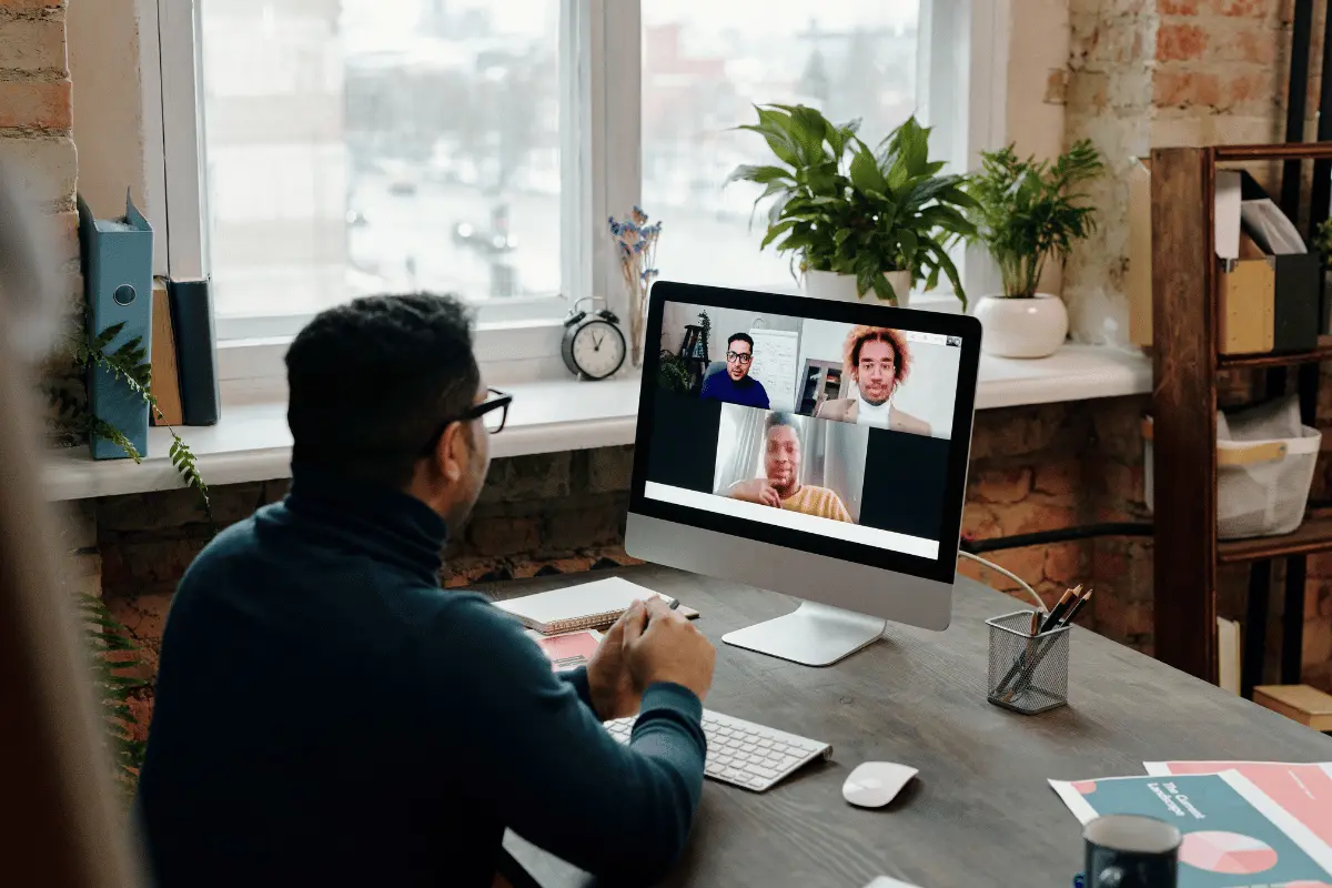 A man at a desk video-calls three colleagues in a bright home office, with leafy plants and a city view creating a calm, collaborative mood.