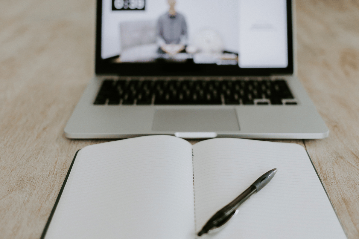 Remote meeting scene with a laptop showing a person on screen and a notebook for note-taking.