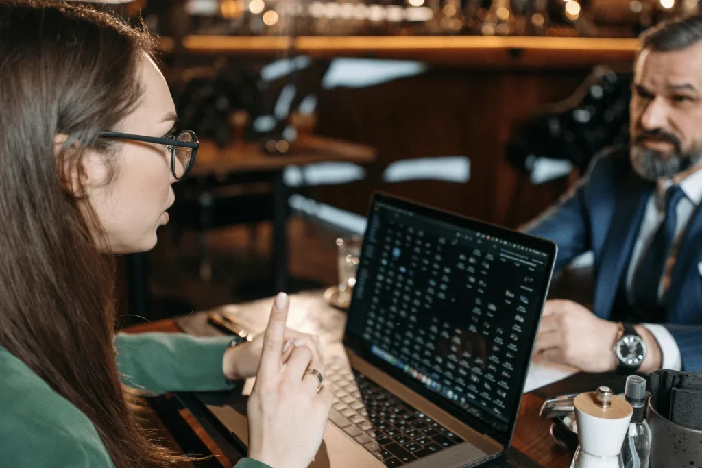 A woman points at a laptop screen while discussing with a man in a business suit at a cafe.