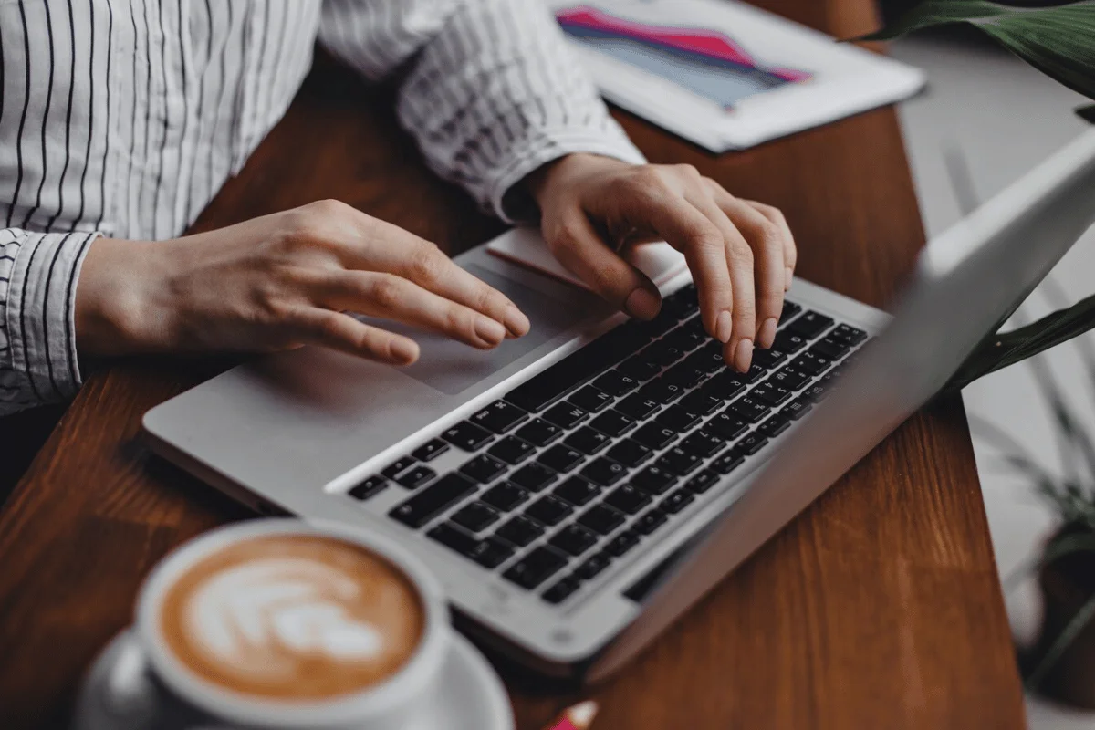 Close-up of hands typing on a laptop with a cappuccino on the side.