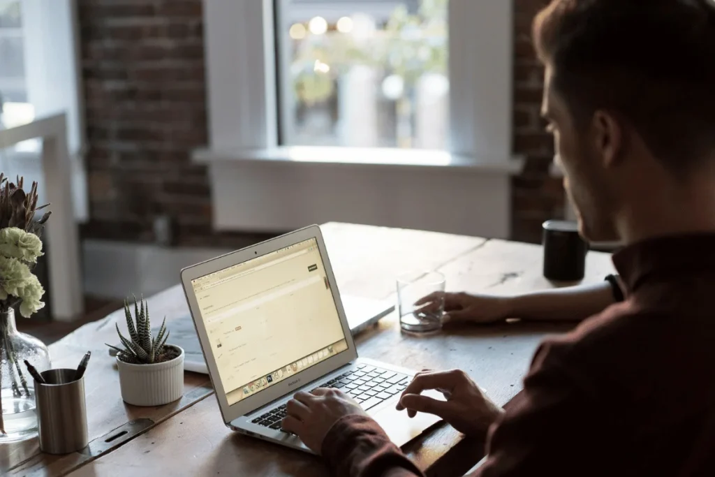 Remote worker typing on a MacBook at a wooden desk near a window.