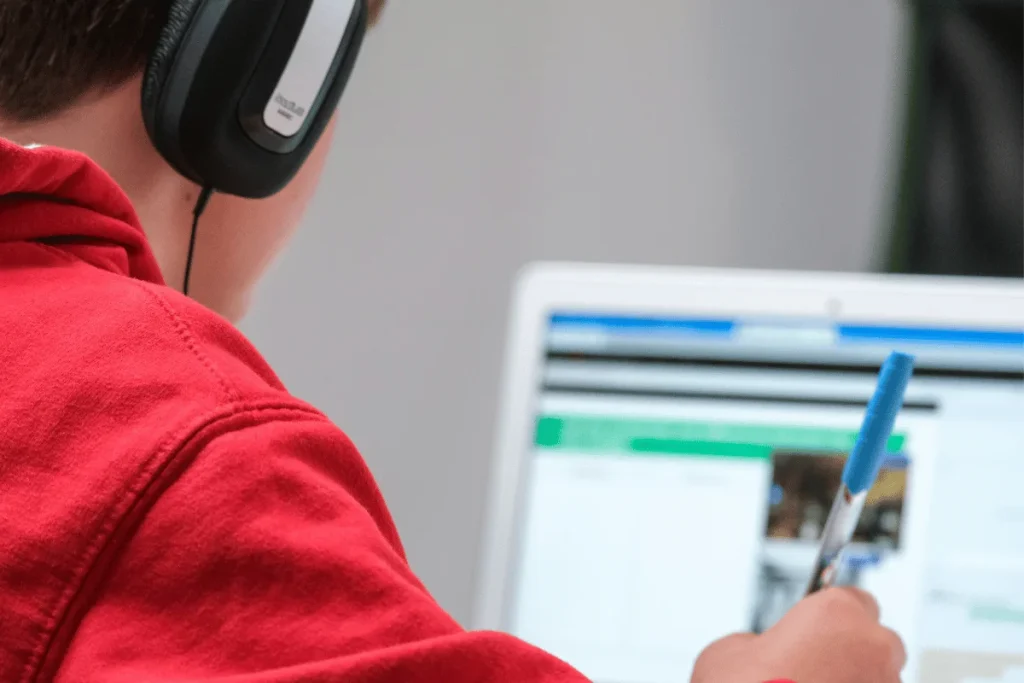Young adult studying on a laptop with headphones and a blue pen in hand.