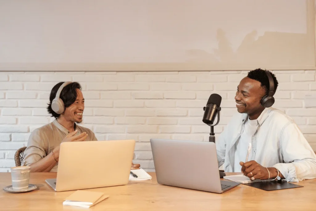 Two podcasters wearing headphones, recording a conversation with laptops and a microphone.