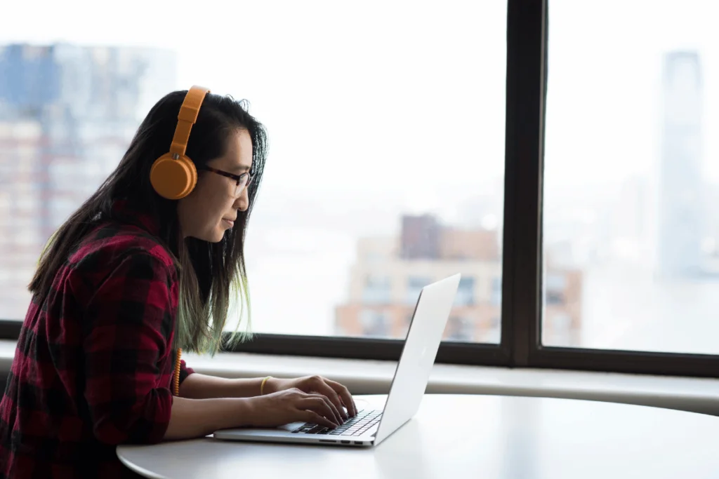 A person wearing orange headphones and glasses is focused on a laptop at a round table, with a cityscape visible through large windows, creating a calm atmosphere.