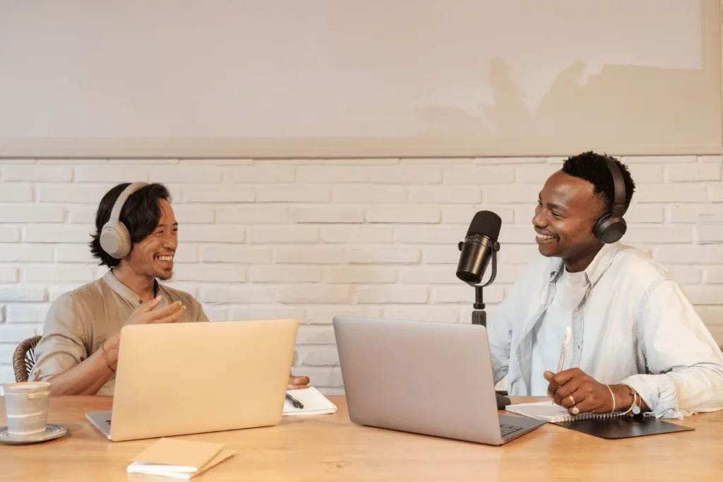 Two podcasters in a studio smiling and recording a podcast with laptops and microphones.