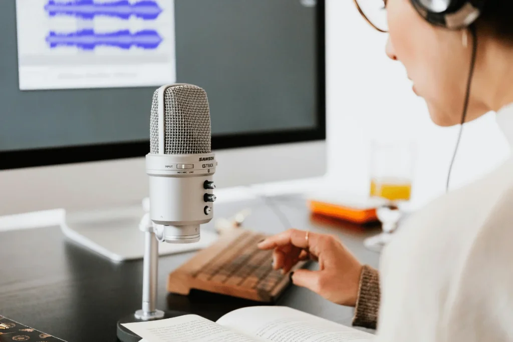 Person recording audio at a desk using a condenser microphone, headphones, and computer with audio waveform on screen.