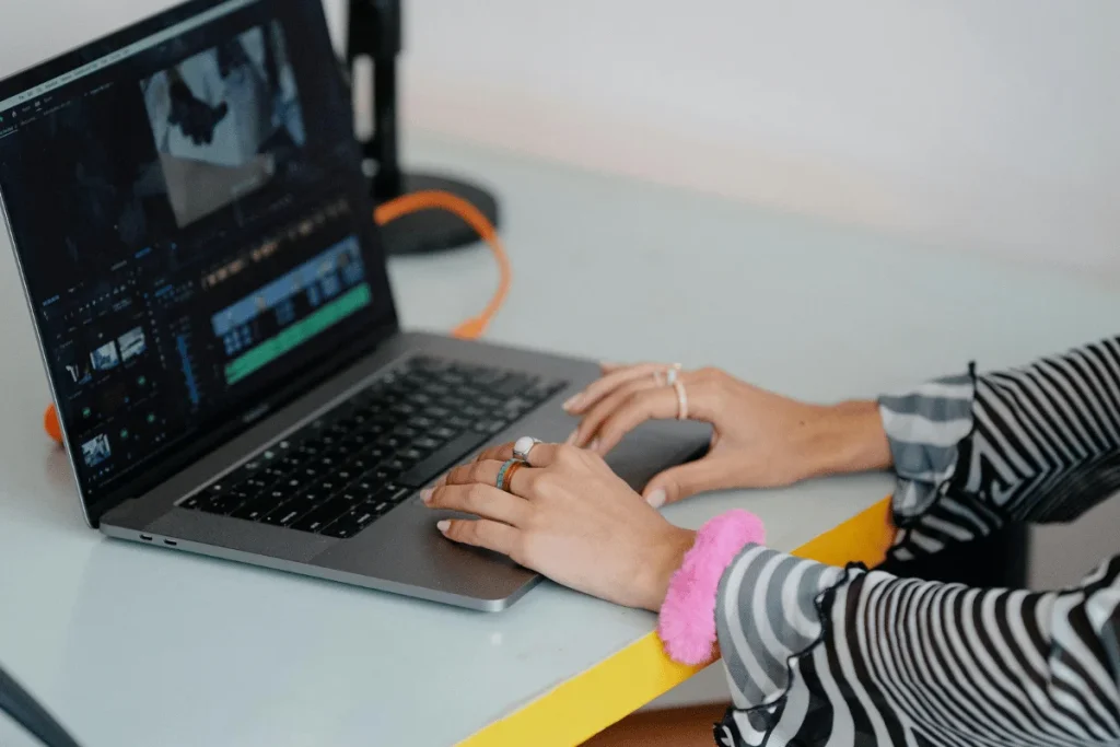 A person edits video on a laptop with colorful rings and a pink scrunchie, focused on the screen's editing software.