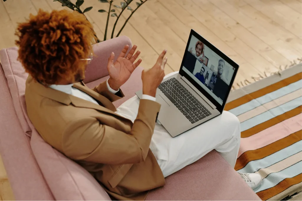Businessperson on a pink sofa engaging in a virtual meeting with four team members visible on a laptop screen.
