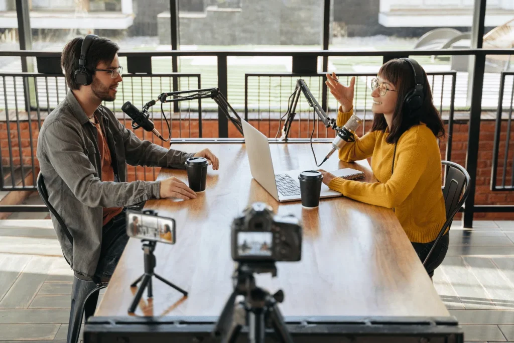 Two podcasters engage in discussion at a wooden table with microphones, laptops, and coffee cups in a bright studio setting.