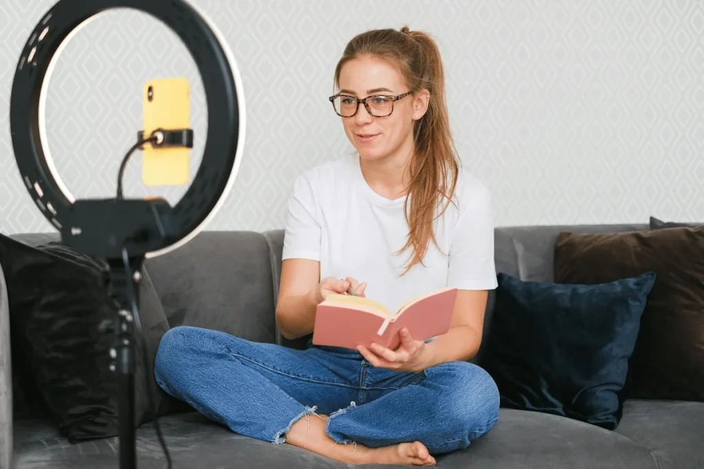Young woman reading a book while recording herself with a smartphone on a ring light in a cozy living room.