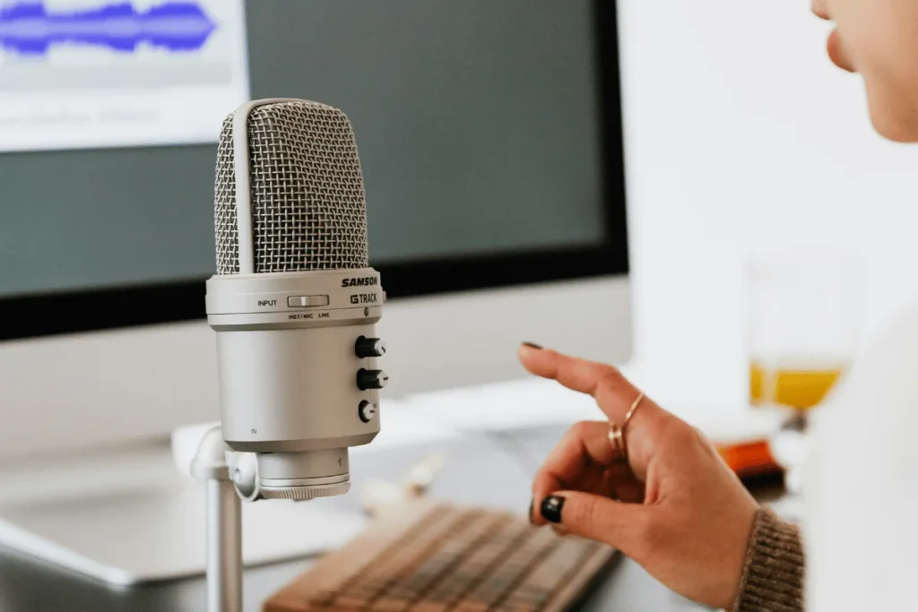 A close-up of a person's hand adjusting settings on a Samson G Track microphone, with a computer screen displaying audio waveforms in the background.