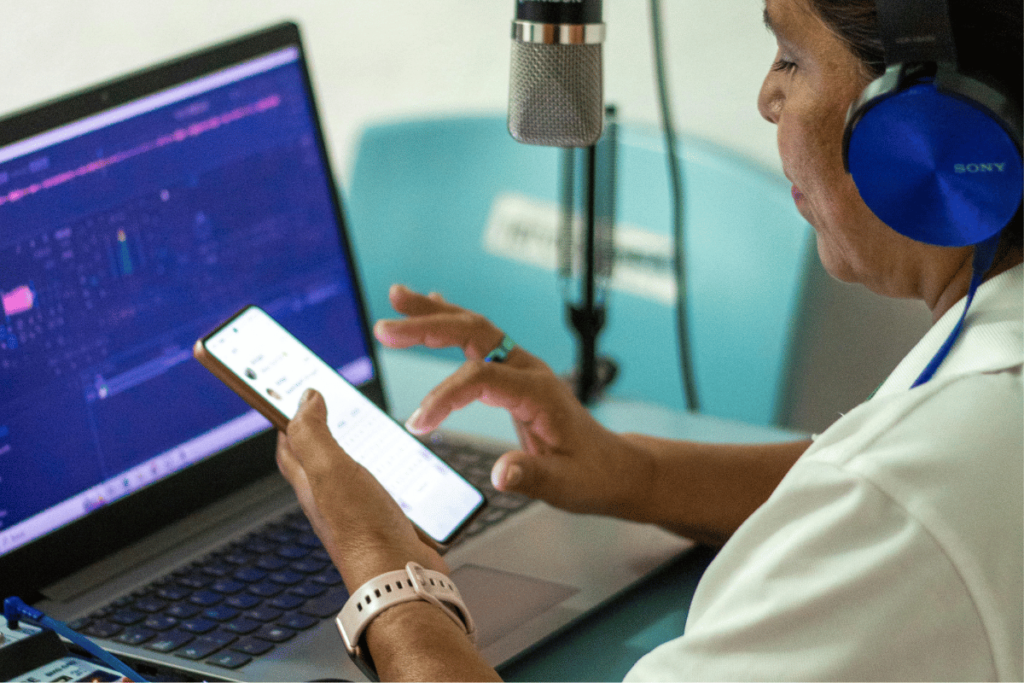 Older woman multitasking with smartphone and laptop in a studio, wearing headphones and a white shirt.