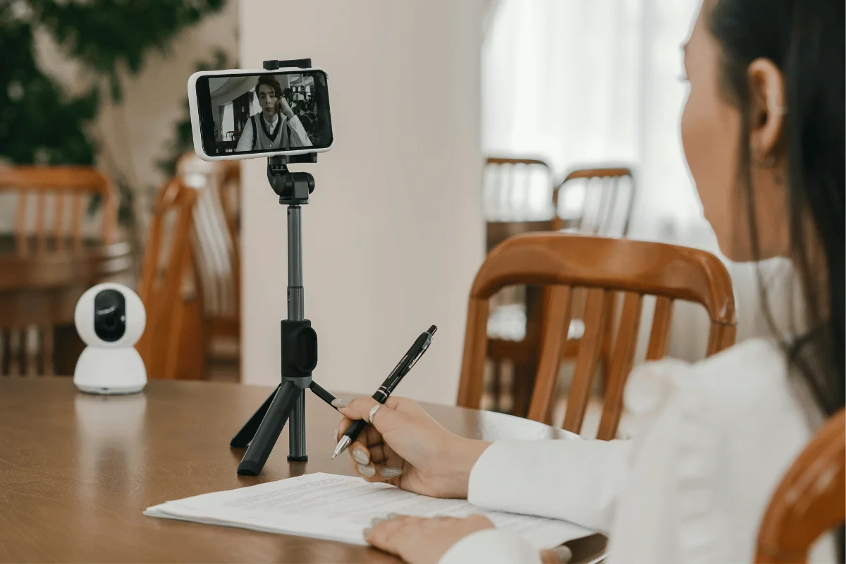Woman participating in an online video meeting using a smartphone on a tripod while taking notes in a well-lit room with wooden chairs.