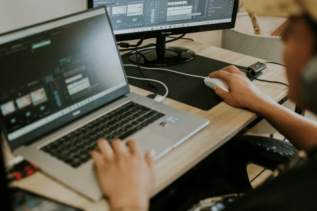 A person editing video on a laptop, using a mouse, with a second monitor displaying editing software tools on a wooden desk.