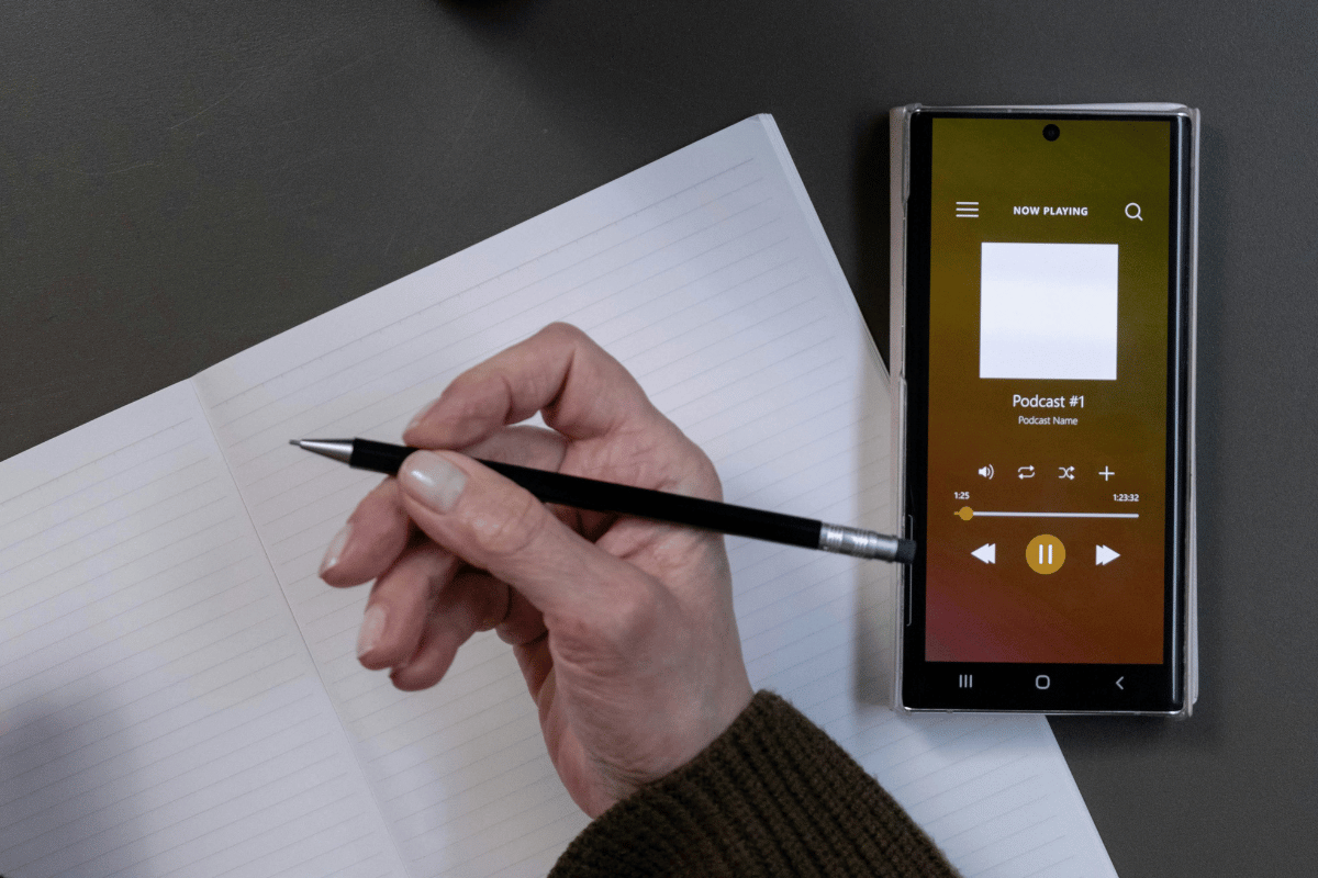 Hand with pen ready to take notes beside a smartphone with a podcast app on the screen.