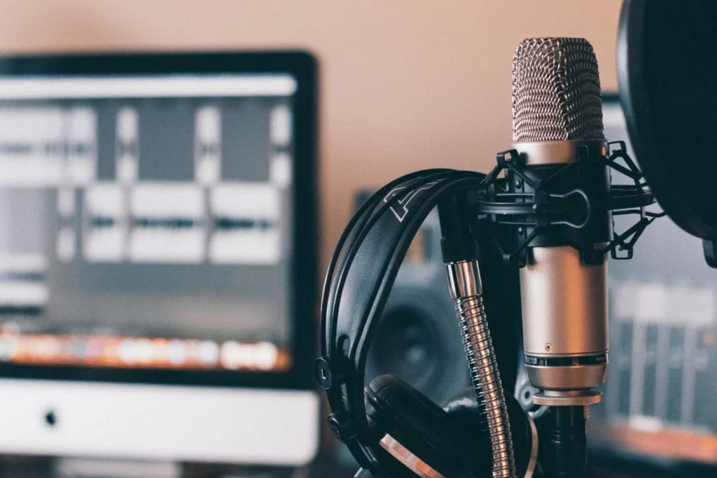 Close-up of a microphone with headphones resting on its stand, against a blurred computer screen displaying audio waveforms.