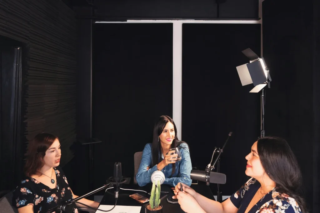 A group of three women engage in conversation at a podcast table, with microphones and soft lighting in a modern studio setting.