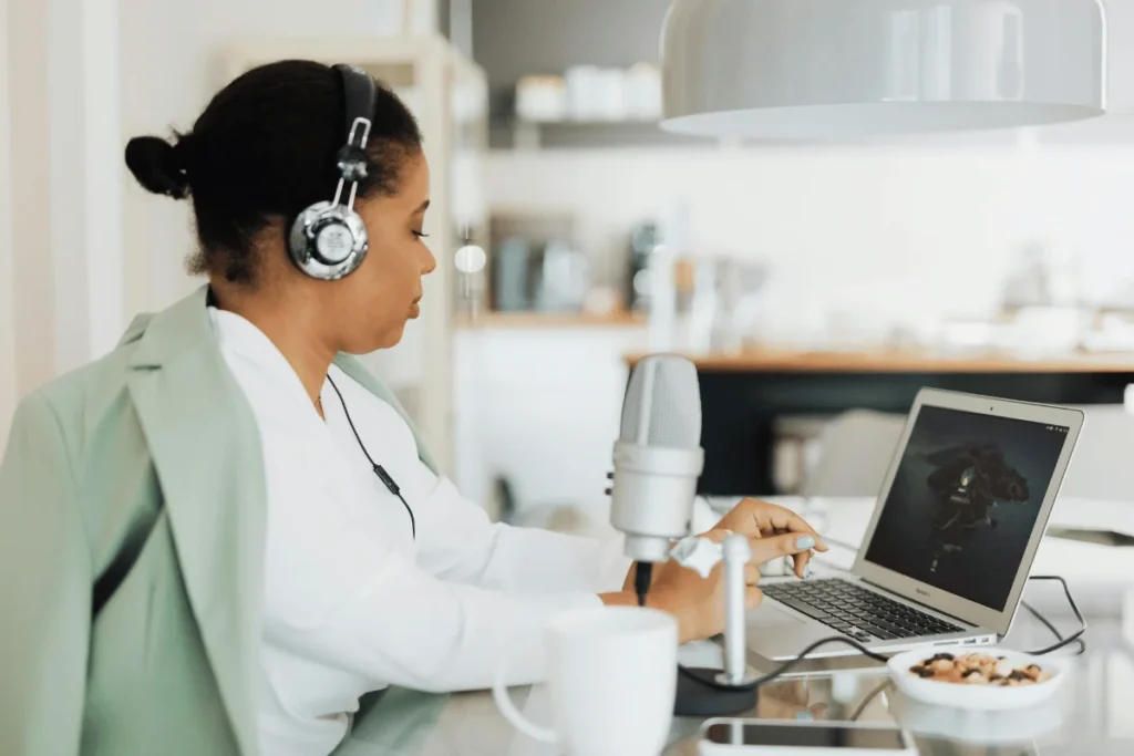 Female podcaster with headphones and microphone, working on a laptop in a modern home office setting.