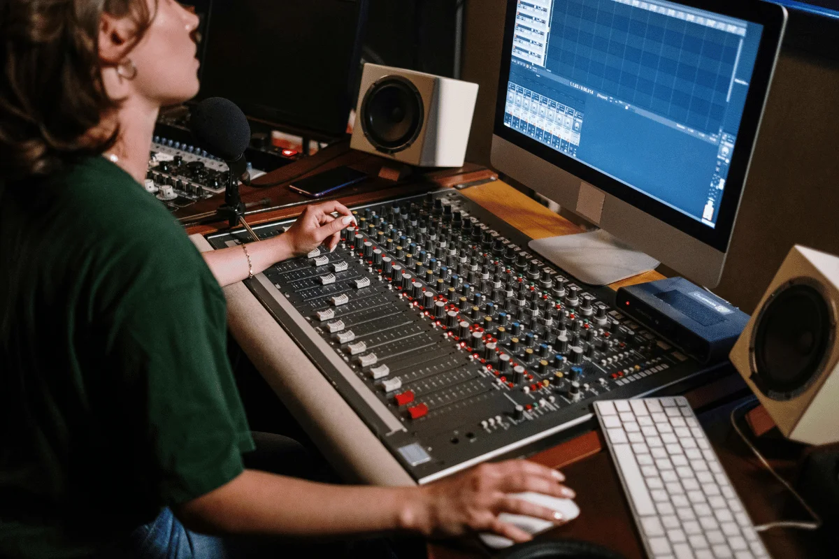 Audio engineer adjusting a mixing console in a studio, with an iMac displaying audio tracks and speakers on the desk.