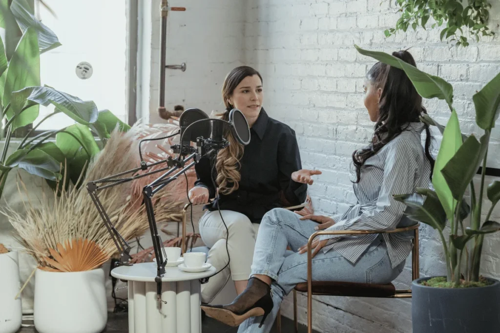 Two women engaged in a podcast recording session, discussing in a well-lit, plant-filled studio.