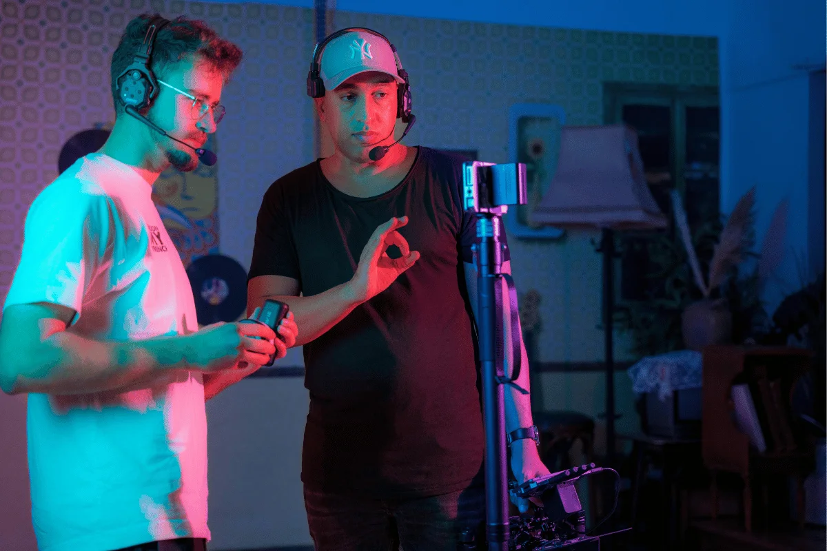 Two men wearing headsets working on a film set, adjusting camera equipment with colorful lighting.