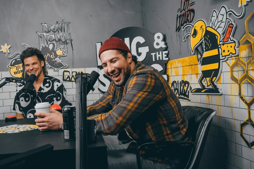 Two podcasters laughing during a lively recording session with colorful graffiti in the background.