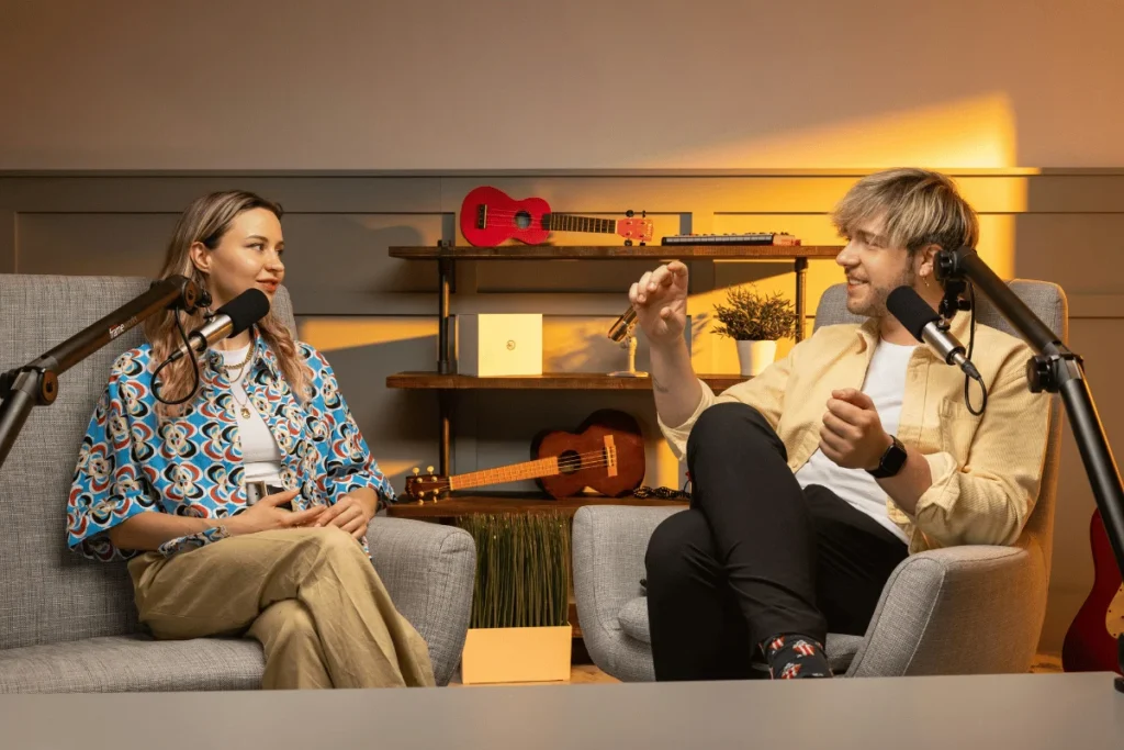 Two podcasters engaged in a lively discussion, sitting comfortably in chairs with microphones.