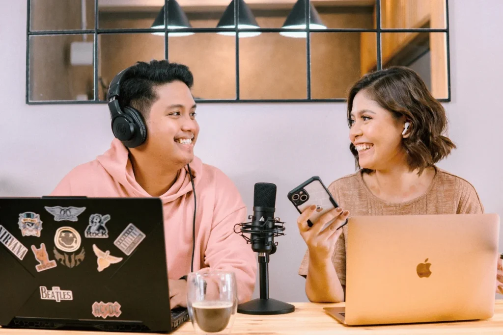 Two people smiling while recording a podcast, each with a laptop and a microphone between them.