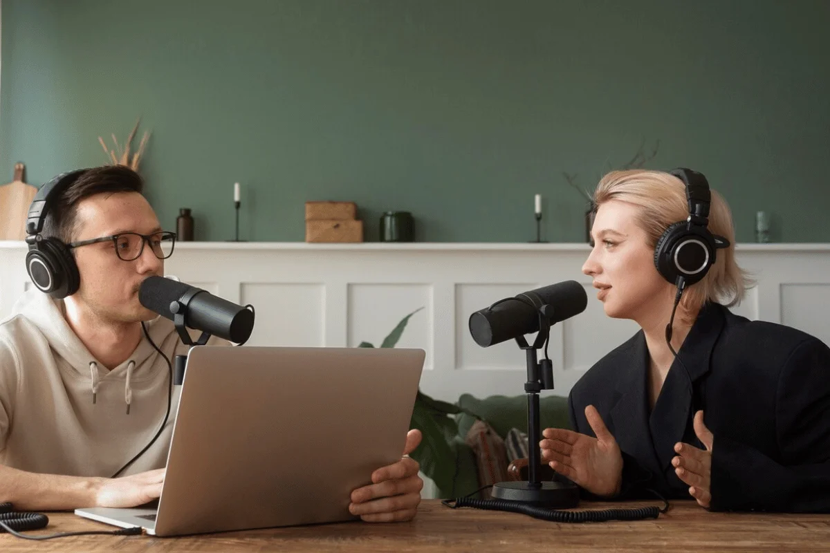 Two podcast hosts, one male and one female, recording an episode in a well-decorated studio with a laptop.