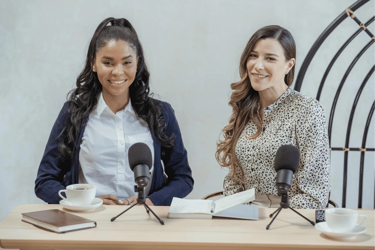 Two female presenters smiling at a table with microphones and coffee cups, preparing for a podcast.