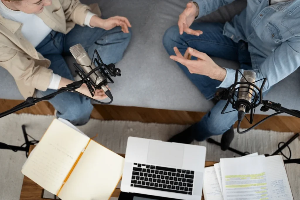 Two podcasters sitting on a couch with microphones and a laptop, discussing content for their show.