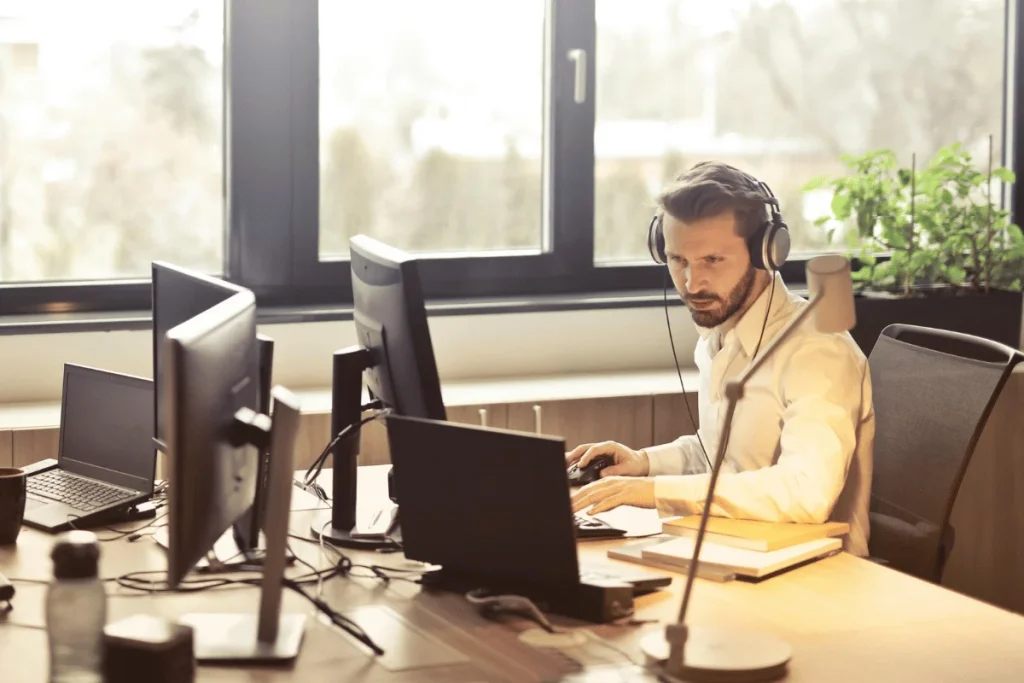 Man in white shirt using computer with headset in a sunlit modern office.