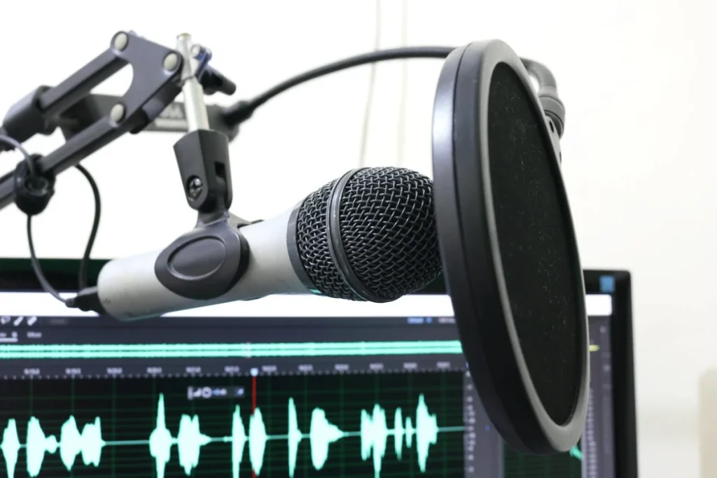 Professional microphone with pop filter and audio waveforms on a computer screen in a recording studio.