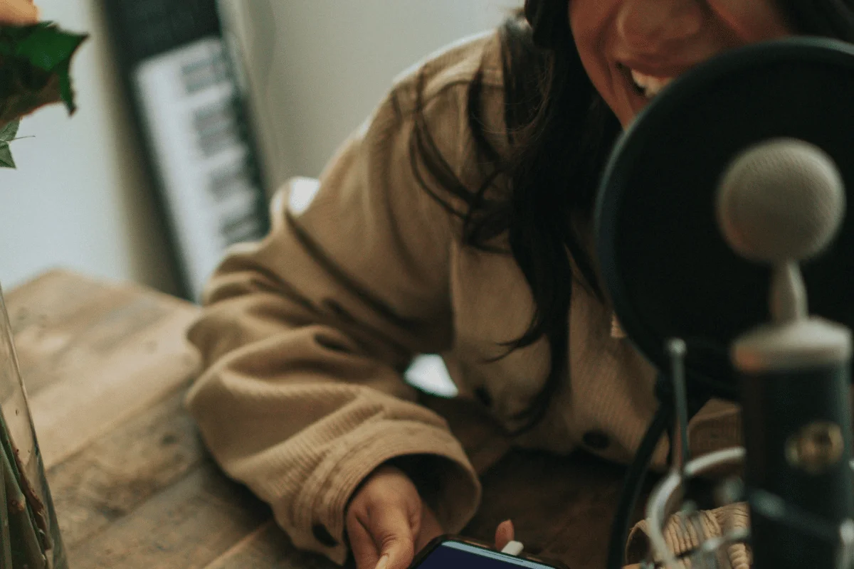 A woman smiles while speaking into a microphone, holding a smartphone, suggesting a recording or podcast session.