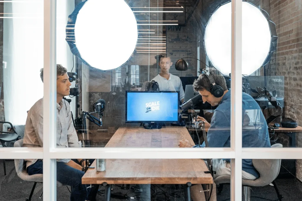 Two men recording a podcast in a studio with professional microphones and lighting setup, viewed through a glass partition.