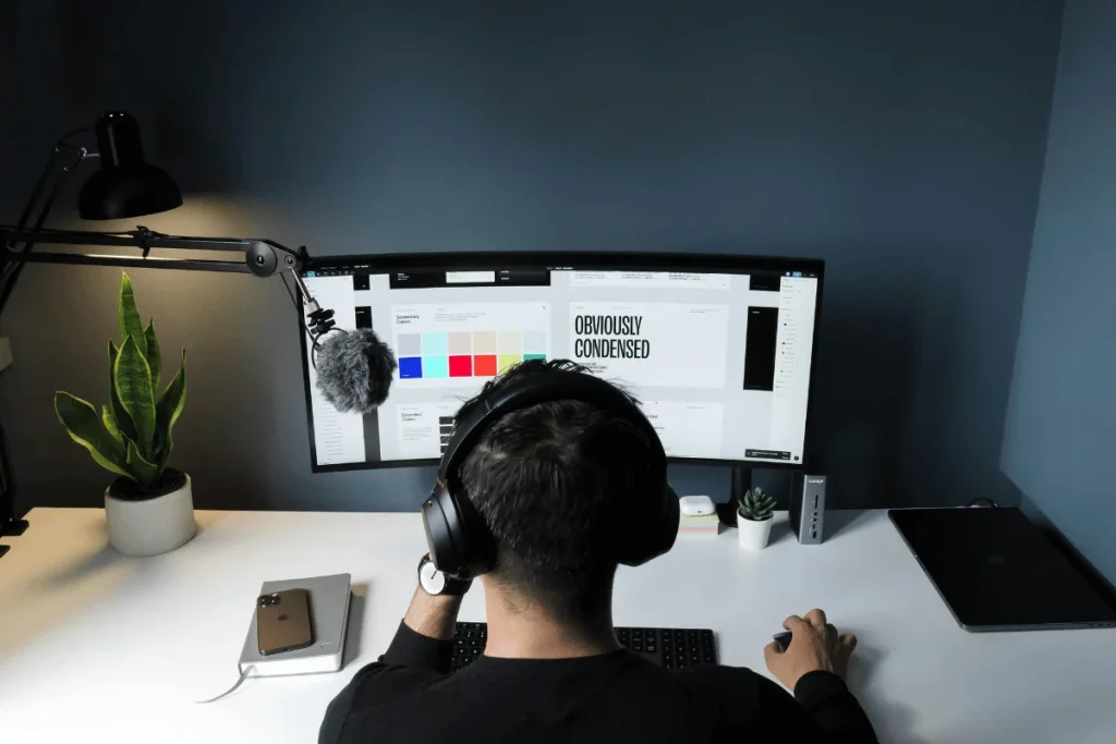 Designer with headphones working on a graphic project on a wide-screen monitor.