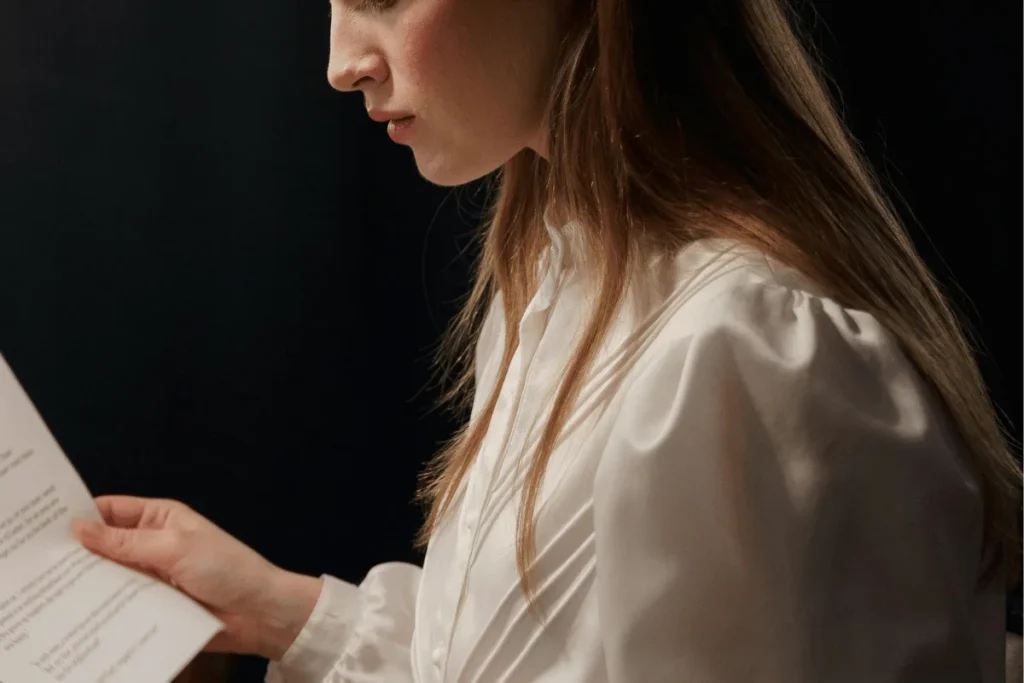 Woman in a white blouse reading a document against a dark background, focused on the text in her hand.