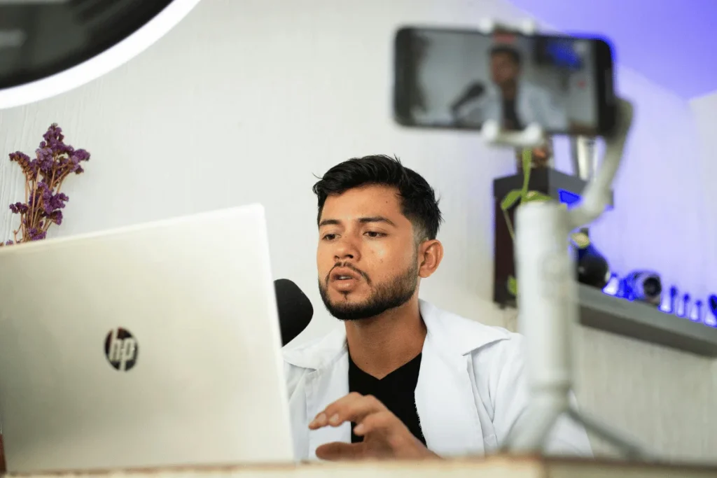 Man speaking into a microphone while working on a laptop with a phone recording setup.