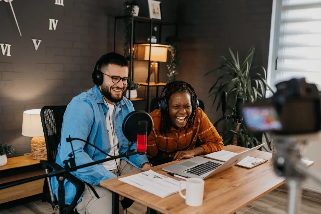 Two people wearing headphones sit at a wooden table with a laptop, microphone, and documents, engaged in a podcast or video recording session.