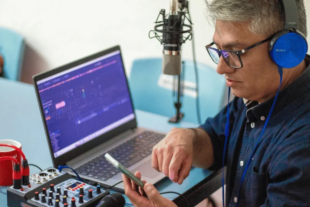 Podcaster checking his phone during a recording session, with a microphone and audio mixer on the table.