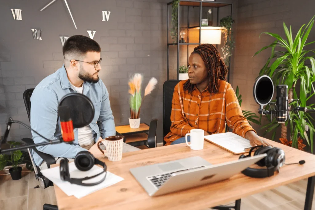 Two people sitting at a wooden desk with microphones, headphones, and a laptop, engaged in a podcast conversation.