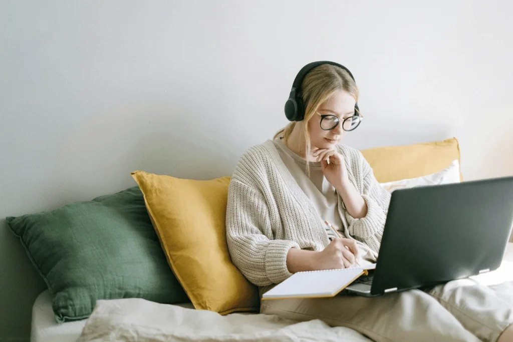 A person wearing headphones sits on a couch, working on a laptop, with a notebook in hand and colorful cushions behind them.