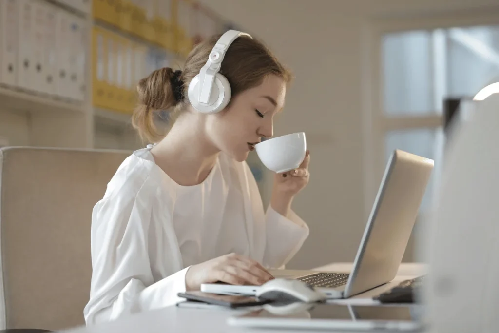 Young woman in white headphones sipping coffee while working on a laptop.