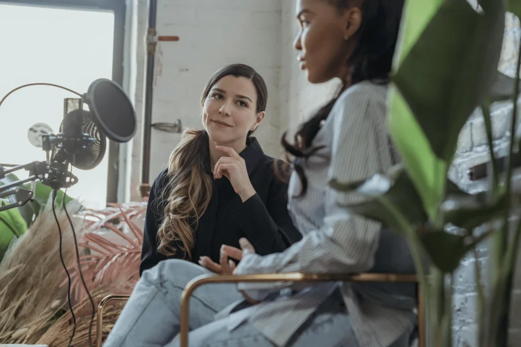 Podcast hosts in discussion, recording in a bright studio with a professional microphone and greenery.