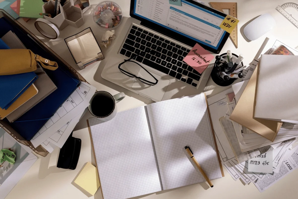 Cluttered desk with laptop, eyeglasses, and assorted office supplies.